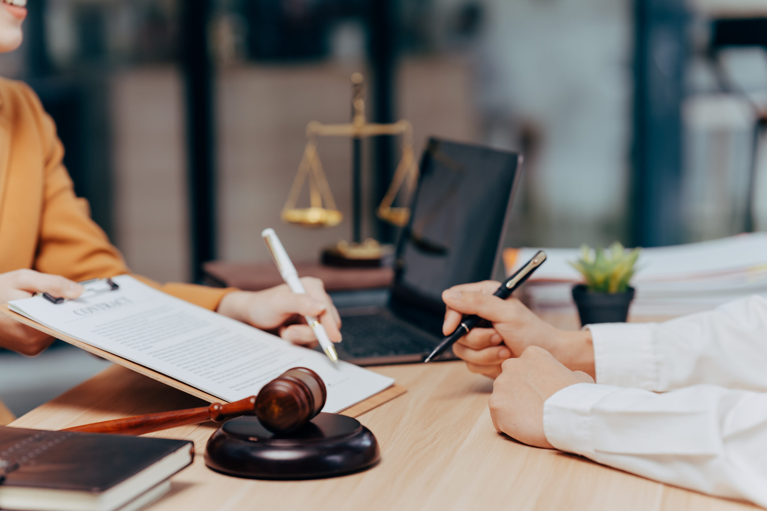 Legal Consultation: Two professionals sit at a desk, carefully reviewing and signing legal documents. In the background a scale of justice rests, and a gavel sits nearby.
